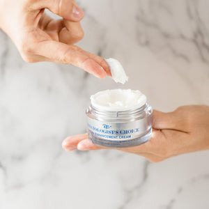 Hand holding an open jar of Facial Enhancement Cream Mild Glycolic Daily Moisturizer | Dermatologist's Choice, with a finger scooping out a small amount of white cream against a light marble background.