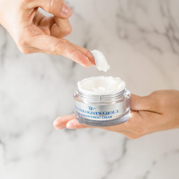 Hand holding an open jar of Facial Enhancement Cream Mild Glycolic Daily Moisturizer | Dermatologist's Choice, with a finger scooping out a small amount of white cream against a light marble background.