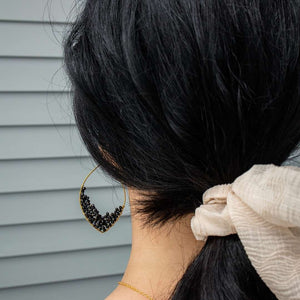 Close-up of a woman's dark hair tied with a light fabric scrunchie, showcasing a delicate gold and black beaded earring from Dusk Drops | Purpose Jewelry.