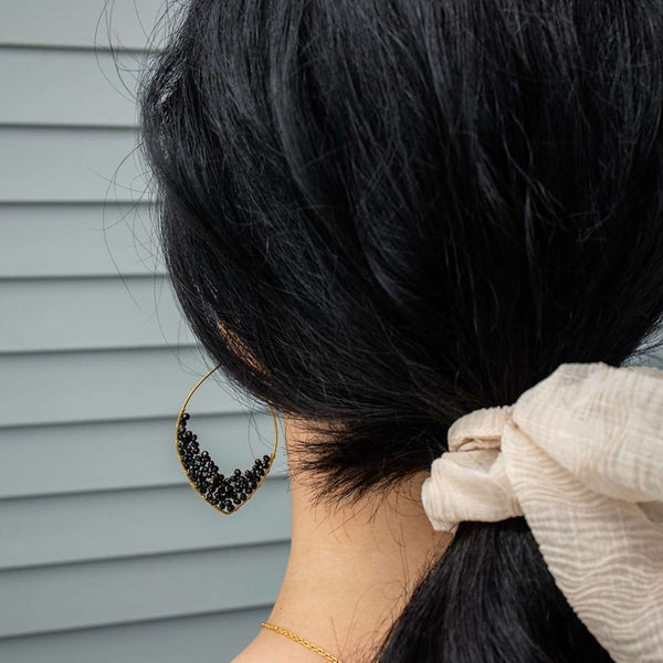 Close-up of a woman's dark hair tied with a light fabric scrunchie, showcasing a delicate gold and black beaded earring from Dusk Drops | Purpose Jewelry.