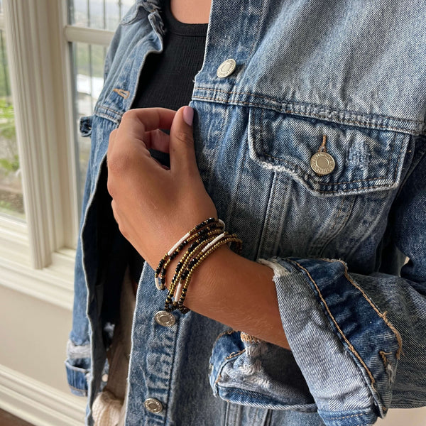 Close-up of a person wearing layered beaded bracelets featuring dark and metallic tones with a charm, styled with a denim jacket and black top, showcasing the Dusk Stretch Bracelet | Purpose Jewelry.