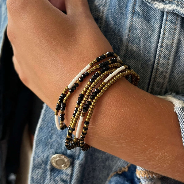Close-up of a wrist wearing multiple beaded strands in black, white, and gold tones styled with denim fabric, showcasing the Dusk Stretch Bracelet | Purpose Jewelry.