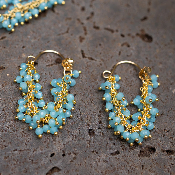 Elegant gold-tone Fringe Hoop Earrings | Catherine Weitzman Jewelry adorned with clustered light blue beads, displayed on a textured surface for a stylish look.