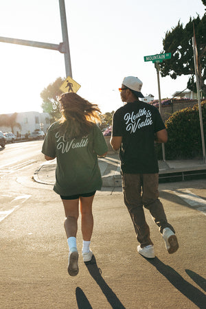 Two people wearing "Health Is Wealth T-Shirt | Lucky Owl" walking on a sunny street, showcasing casual wellness-inspired apparel in an urban setting.
