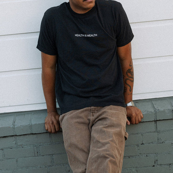 Man wearing a black Health Is Wealth T-Shirt | Lucky Owl sitting casually against a gray painted brick wall, paired with light brown pants and a wristwatch.