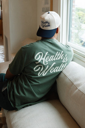 Person wearing a green "Health Is Wealth T-Shirt | Lucky Owl" sitting on a beige couch by a window, with a matching cap also featuring the "Health is Wealth" text.