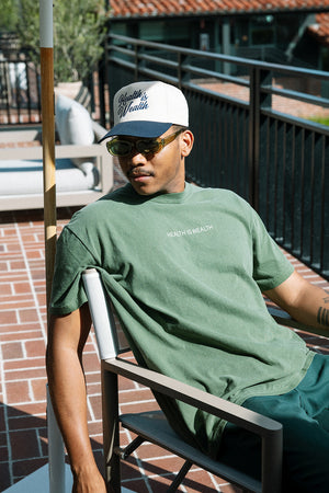 Man wearing sunglasses and a white and black cap sitting outdoors on a patio chair, dressed in a green Health Is Wealth T-Shirt | Lucky Owl, relaxing in sunlight.