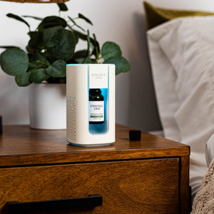 Essential Oil Blends | Essence One diffuser with a blue essential oil bottle inside, placed on a wooden bedside table next to a green plant and white bedding, promoting skincare and wellness.