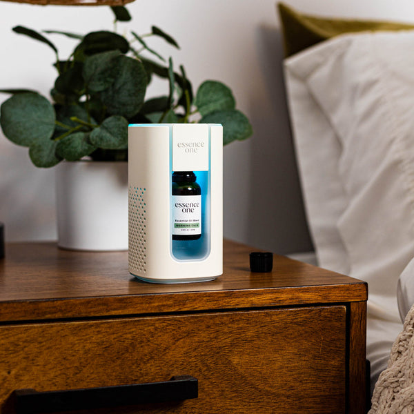 Essential Oil Blends | Essence One diffuser with a blue essential oil bottle inside, placed on a wooden bedside table next to a green plant and white bedding, promoting skincare and wellness.