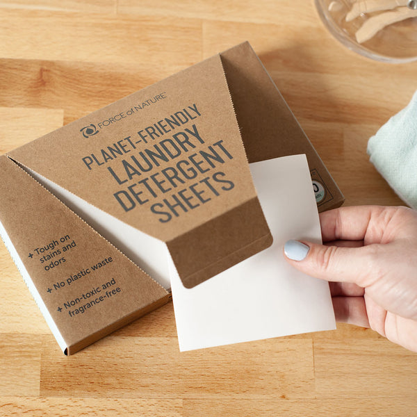 Hand holding a white laundry detergent sheet being pulled from an open box of Laundry Detergent Sheets | Force of Nature on a wooden surface.