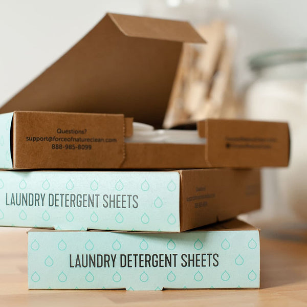 Stack of eco-friendly laundry detergent sheets boxes with one open showing white sheets inside, promoting Laundry Detergent Sheets | Force of Nature in a clean, minimalist setting.