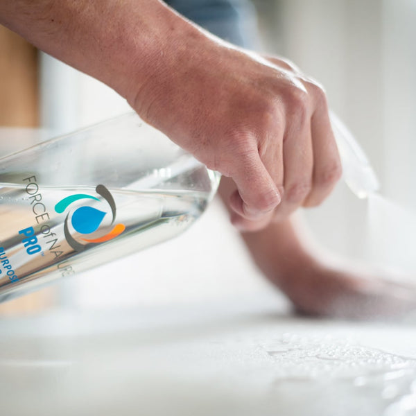 Hand pouring clear liquid from a Force of Nature Pro spray bottle onto a white surface, highlighting Activator Capsules 25ct box 12oz/3mL | Force of Nature for wellness and body care use.