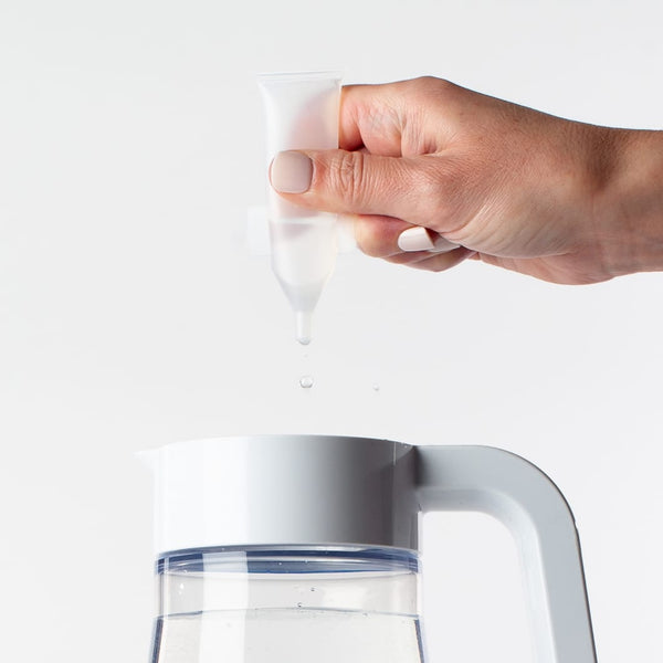 Hand holding a small clear capsule dripping liquid into a white water pitcher, Activator Capsules 25ct box 12oz/3mL | Force of Nature.