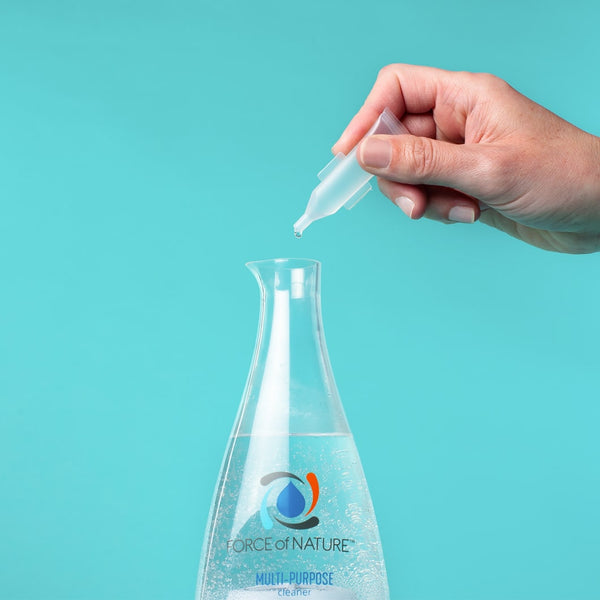 Hand holding a dropper bottle adding liquid into a clear glass bottle filled with solution labeled Force of Nature, 12oz Glass Bottle Cleaning Starter Kit | Force of Nature on a teal background
