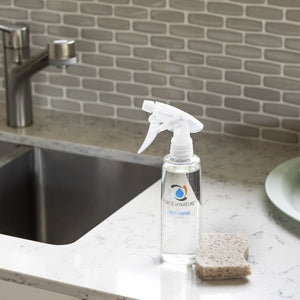 Clear spray bottle labeled 12oz Glass Bottle Cleaning Starter Kit | Force of Nature on a kitchen counter beside a sponge near a stainless steel sink and faucet.