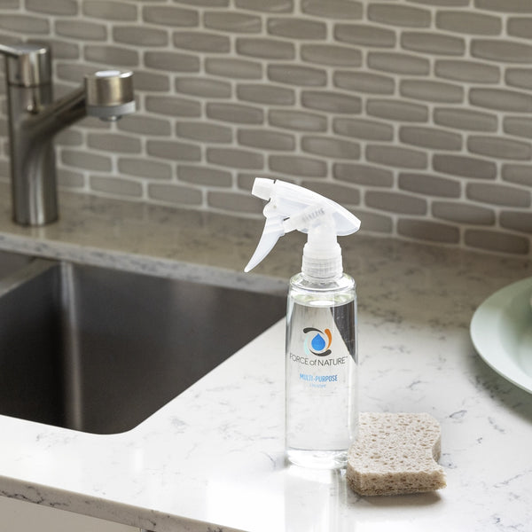 Clear spray bottle labeled 12oz Glass Bottle Cleaning Starter Kit | Force of Nature on a kitchen counter beside a sponge near a stainless steel sink and faucet.