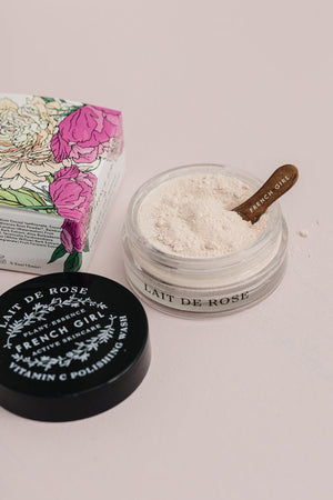 Open jar of Vitamin C Polishing Facial Wash & Mask | French Girl with a wooden spatula resting inside, surrounded by the product’s floral packaging and black lid on a pale surface.