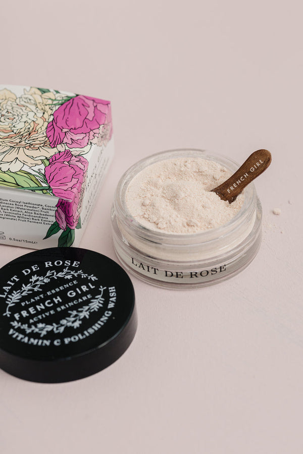 Open jar of Vitamin C Polishing Facial Wash & Mask | French Girl with a wooden spatula resting inside, surrounded by the product’s floral packaging and black lid on a pale surface.