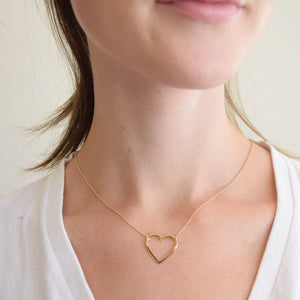 Close-up of a woman wearing a delicate gold heart-shaped pendant necklace, the "Follow Your Heart Silhouette Necklace (Small / Gold) | Love You More," styled with a white top.