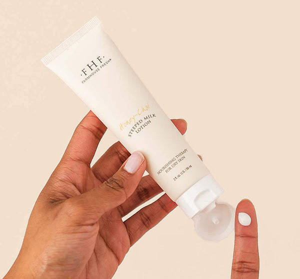 Hand holding a tube of Honey-Chai Steeped Milk Lotion® for Hands | Farmhouse Fresh with a small amount of lotion on a fingertip against a beige background, highlighting skincare and body care.