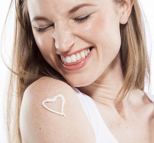 A smiling woman with light skin applying a heart-shaped dollop of Honey-Chai Steeped Milk Lotion® for Hands | Farmhouse Fresh on her shoulder, promoting skincare and body care.