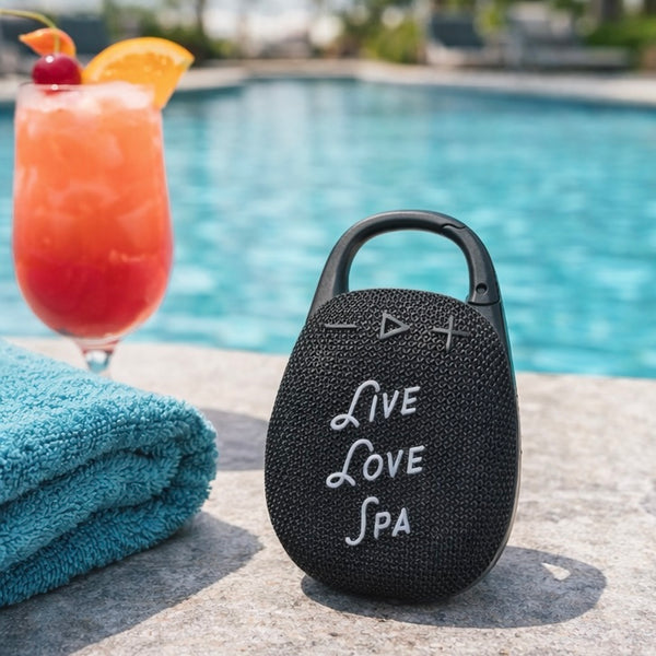 Wireless Clip Speaker | Live Love Spa in black with textured surface, placed by a pool next to a blue towel and a tropical drink, showcasing a portable wellness device for outdoor use.
