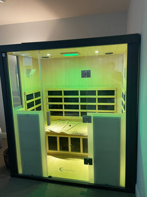 Modern wooden sauna cabin with glass doors, illuminated by soft green light, featuring a bench with towels inside for relaxation and wellness using HaloSauna™ | Halotherapy Solutions.