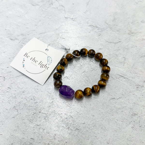 Tiger's Eye Luxe Bracelet | Be the Light featuring polished brown tiger's eye beads with a single purple stone accent, displayed on a light textured surface with a branded tag.