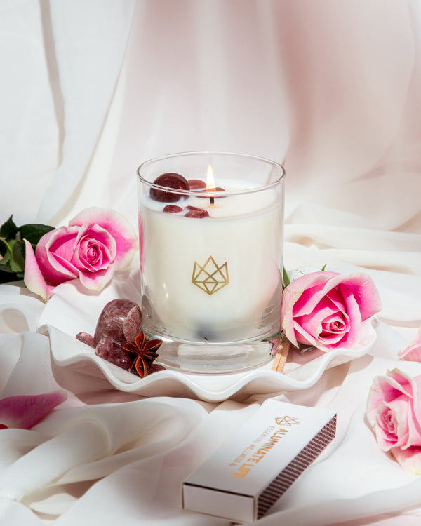 Sensual Glass Candle | Aluminate Life burning softly in a clear glass holder with decorative stones, surrounded by pink roses and a branded matchbox on white fabric.