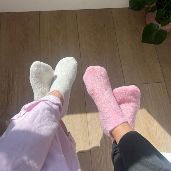Two pairs of feet wearing cozy white and pink socks resting on a wooden floor with sunlight and a green plant nearby, evoking comfort and relaxation with Sip Back & Relax | Barefoot Scientist.