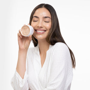 Smiling woman with long dark hair wearing a white robe holds a jar of Kantic® Brightening Moisture Mask | Alchimie Forever close to her face, with some product applied to her cheeks and forehead.