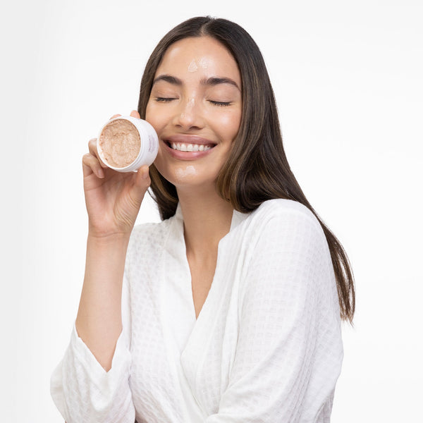 Smiling woman with long dark hair wearing a white robe holds a jar of Kantic® Brightening Moisture Mask | Alchimie Forever close to her face, with some product applied to her cheeks and forehead.