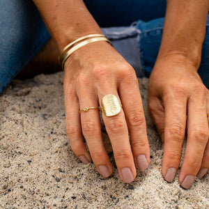 Close-up of hands resting on sand wearing a textured gold ring with a knot detail, two gold bangles, and nude nail polish, featuring the Knot Ring | Purpose Jewelry.