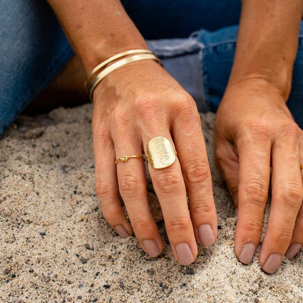 Close-up of hands resting on sand wearing a textured gold ring with a knot detail, two gold bangles, and nude nail polish, featuring the Knot Ring | Purpose Jewelry.