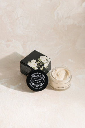 Open jar and closed container of Body Firming Cream | French Girl with floral design on black box and cream texture visible against a light textured background.