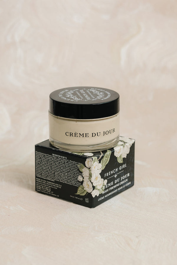 Glass jar of Crème du Jour body firming cream with black lid placed on its floral-printed box on a soft beige surface, showcasing Body Firming Cream | French Girl skincare product.