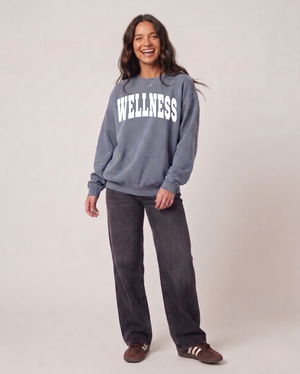 Smiling woman wearing a gray Wellness Unisex Crew Neck Sweatshirt | Lucky Owl paired with dark pants and casual sneakers, standing against a neutral background.