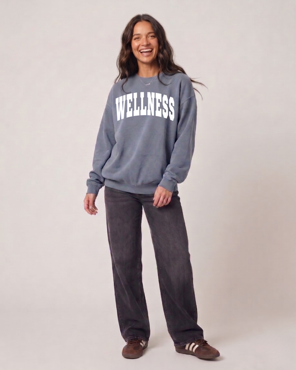 Smiling woman wearing a gray Wellness Unisex Crew Neck Sweatshirt | Lucky Owl paired with dark pants and casual sneakers, standing against a neutral background.