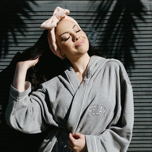 Woman wearing a cozy gray hooded robe with "RELAX" embroidered on the chest, smiling with eyes closed while enjoying sunlight, styled with a soft pink headband, showcasing Limited Edition RELAX Collegiate Robes | Lucky Owl.