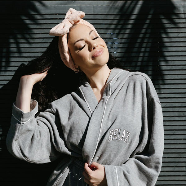 Woman wearing a cozy gray hooded robe with "RELAX" embroidered on the chest, smiling with eyes closed while enjoying sunlight, styled with a soft pink headband, showcasing Limited Edition RELAX Collegiate Robes | Lucky Owl.