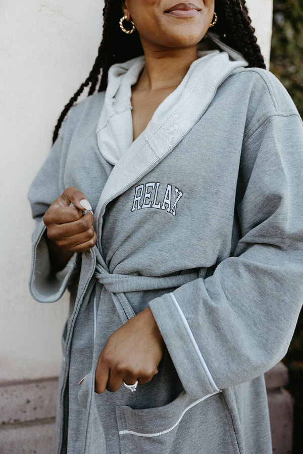 Close-up of a person wearing a cozy grey robe with a hood and "RELAX" embroidered on the chest, featuring a belt and pockets, showcasing the Limited Edition RELAX Collegiate Robes | Lucky Owl.