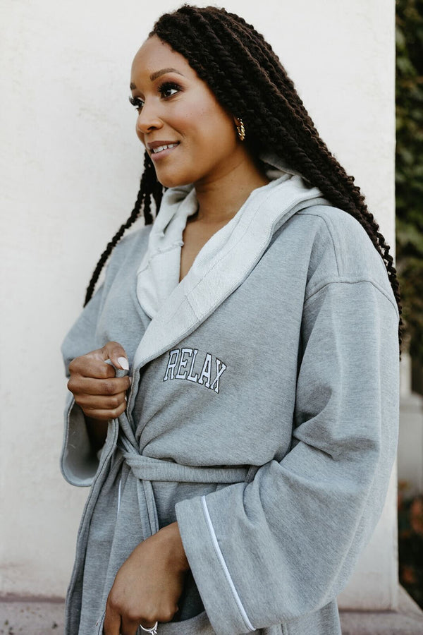 Woman wearing a cozy gray Limited Edition RELAX Collegiate Robes | Lucky Owl with a hood and tie belt, smiling gently while holding the robe closed outdoors.
