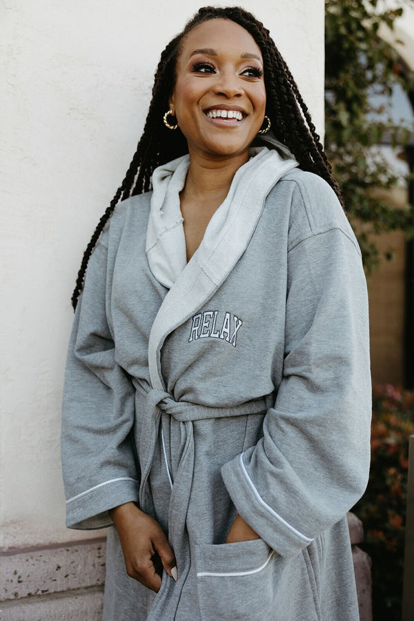 Smiling woman wearing a cozy grey robe with a hood and belt, featuring "RELAX" embroidered on the chest, promoting the Limited Edition RELAX Collegiate Robes | Lucky Owl.