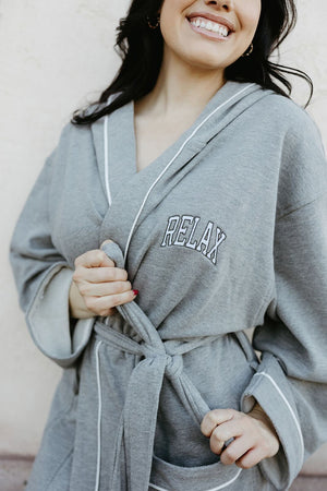 Woman smiling and wearing a soft gray robe with white trim, embroidered with the word RELAX, tied at the waist, showcasing the Limited Edition RELAX Collegiate Robes | Lucky Owl design.