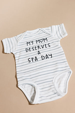 White and light gray striped baby onesie laid flat with black text saying My Mom Deserves A Spa Day, featuring the product name Mom Deserves A Spa Day Baby Onesie | Lucky Owl in a soft, casual style.