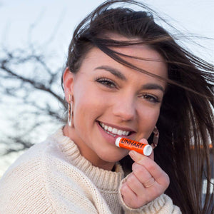 Smiling woman outdoors holding Lip Balm SPF 15 | LINHART near her lips, wearing a cozy cream sweater with wind blowing through her hair, showcasing skincare product use.