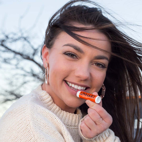 Smiling woman outdoors holding Lip Balm SPF 15 | LINHART near her lips, wearing a cozy cream sweater with wind blowing through her hair, showcasing skincare product use.
