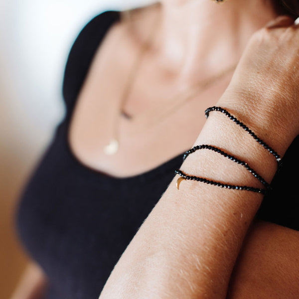 Close-up of a person wearing a black beaded wrap bracelet with a small crescent charm on their wrist, showcasing the Dusk Stretch Bracelet | Purpose Jewelry.