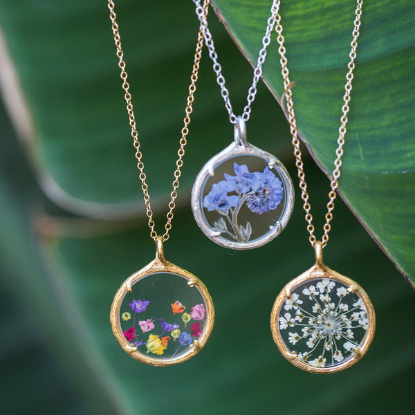 Three Small Botanical Necklace | Catherine Weitzman Jewelry pieces with delicate pressed flowers encased in round glass pendants, hanging on gold and silver chains against green leaves.