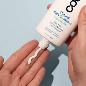 Close-up of a hand dispensing light lotion from a tube of Mineral Body Organic Sunscreen Lotion SPF 50 - Fragrance Free | COOLA on a light blue background, highlighting skincare and body care.